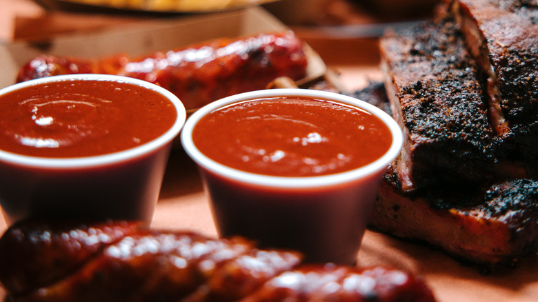 Two small plastic cups of BBQ sauce beside cooked ribs