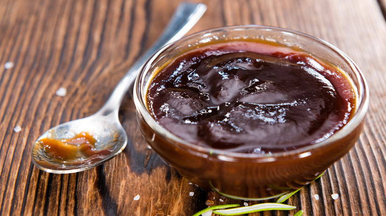A small glass bowl of BBQ sauce with a metal spoon beside it.
