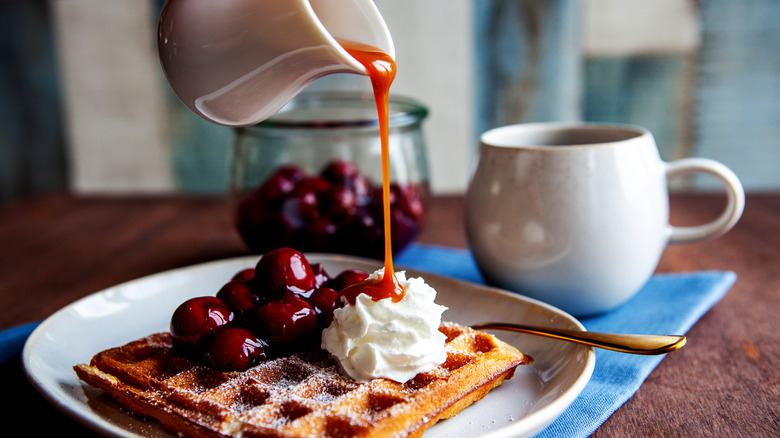 A thick sauce being poured over a waffle with powdered sugar, candied cherries, and whipped cream