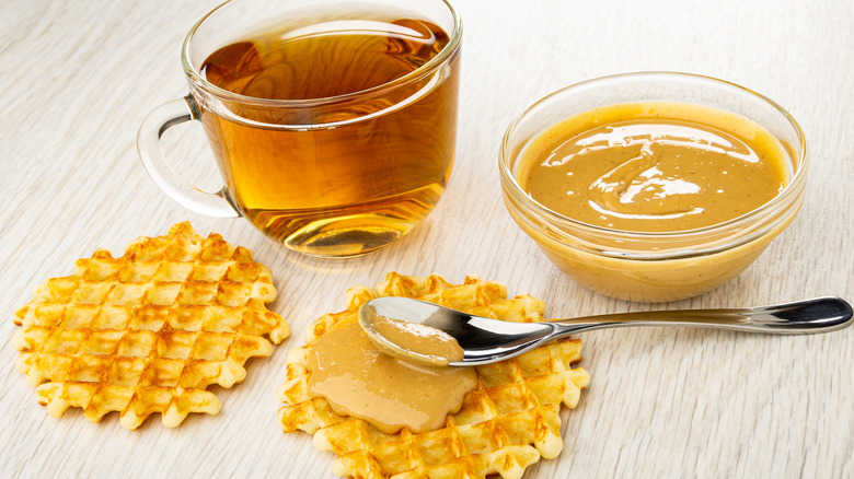 A cup of tea next to a bowl of peanut butter sauce, and two waffles with sauce spread onto one