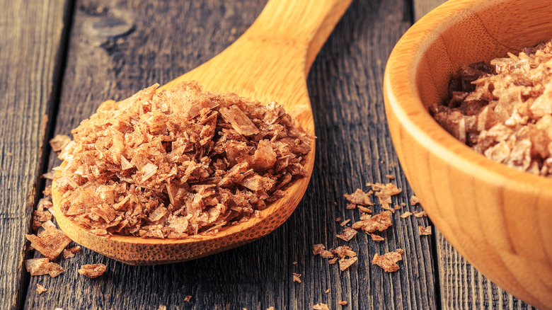 Brown flaky sea salt in a light wooden spoon on a dark wood surface, next to a small wooden bowl of it