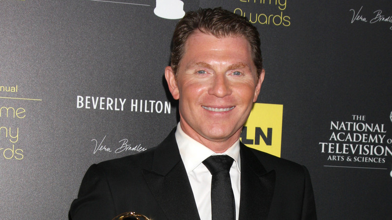 Close-up of Bobby Flay in a black suit with black tie in front of promotional background