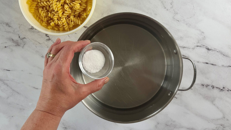 Hand holding small bowl of salt over pot of water