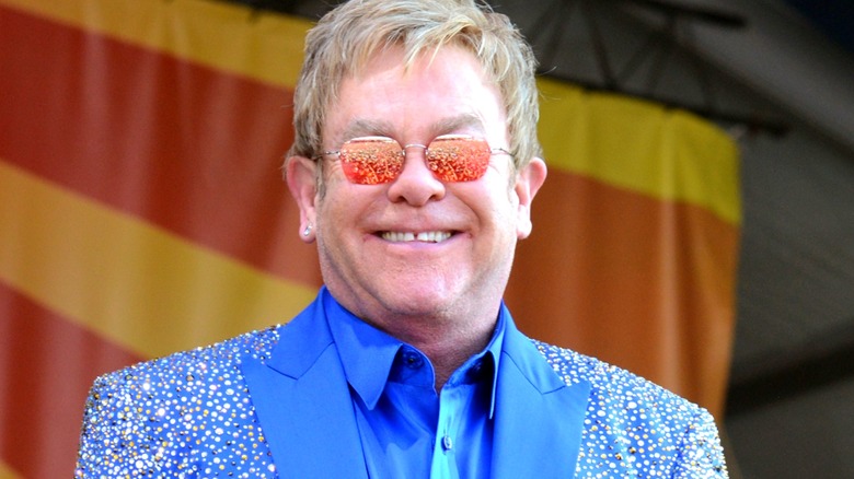 Elton John smiles on stage in orange sunglasses and a blue sparkly suit