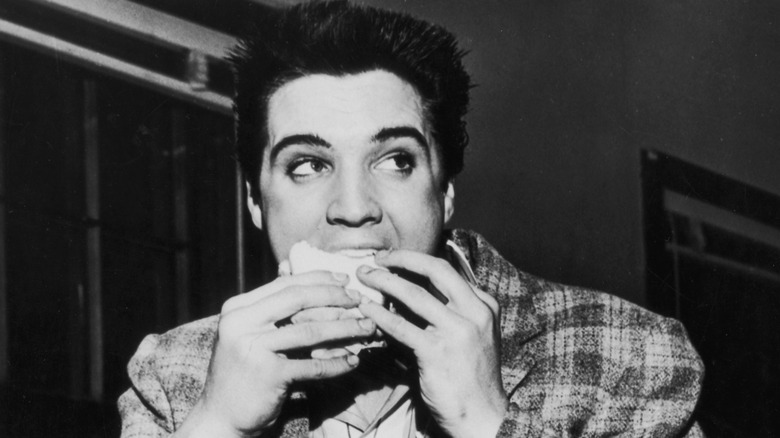 Black and white photo of Elvis Presley taking a bite of a sandwich or pastry