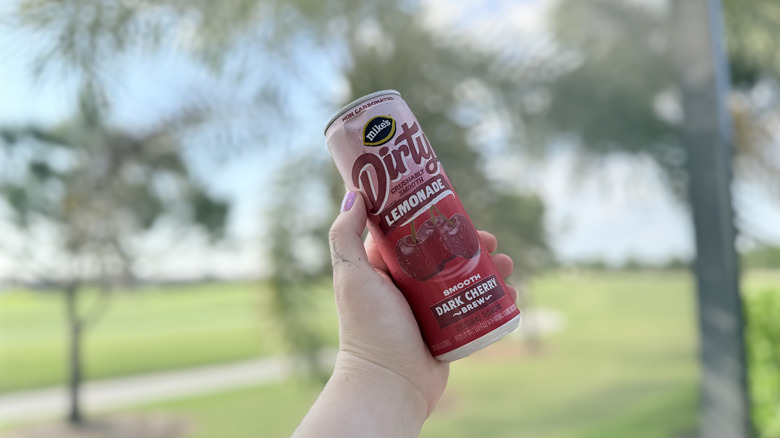 Hand holding a can of Mike's Dirty Lemonade in Dark Cherry Brew flavor with trees and grass in the background