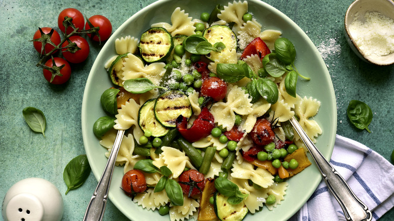 A plate of pasta primavera on a green table surrounded by spinach, tomatoes, and parmesan