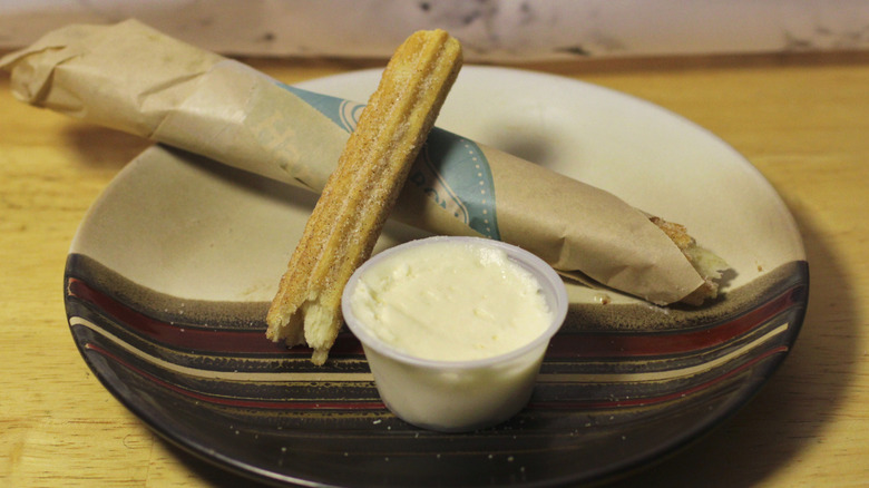 A Cinnabon Churro Stick in two pieces resting on a plate with a side of cream cheese icing