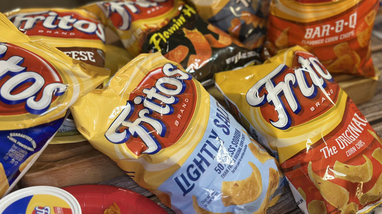 Bags of different flavored Fritos on a wooden surface, including lightly salted, original, and bar b q flavors