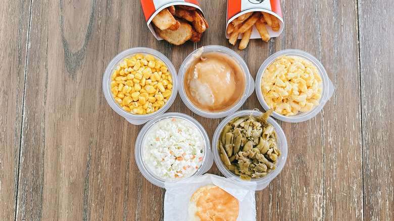 KFC sides arranged on a wooden table, including mashed potato with gravy, corn, and coleslaw