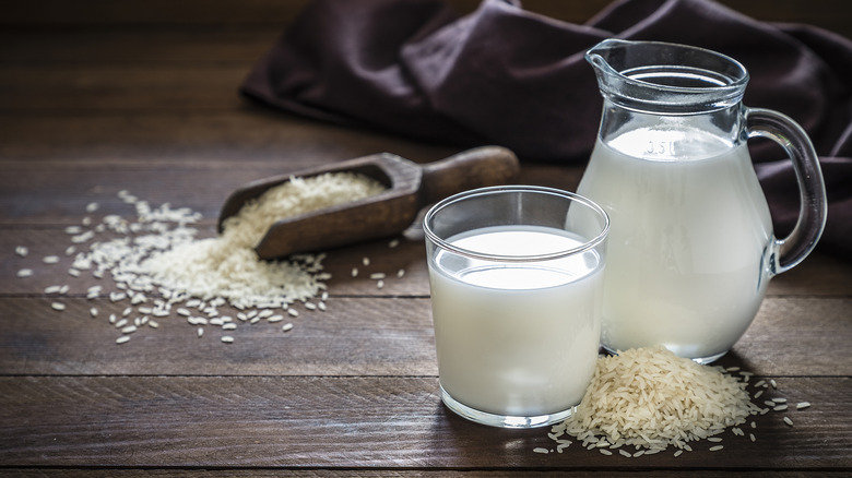 Milk in glass pitcher surrounded by dry rice on brown wooden table