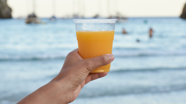 A hand holding plastic cup of orange juice in front of cove