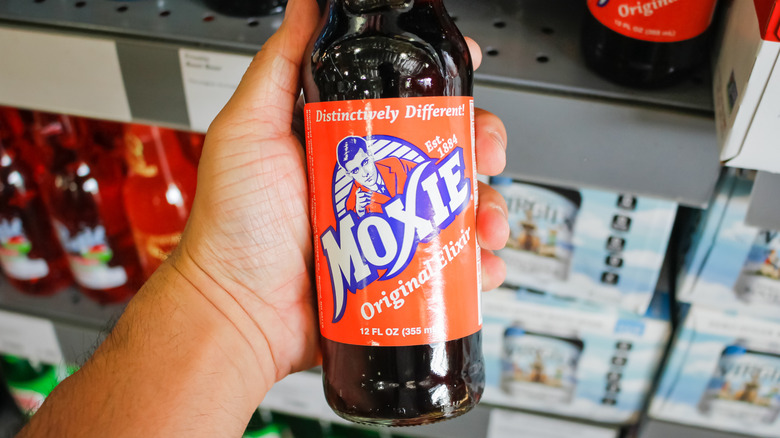 A hand holding a bottle of Moxie in the soda aisle in a store