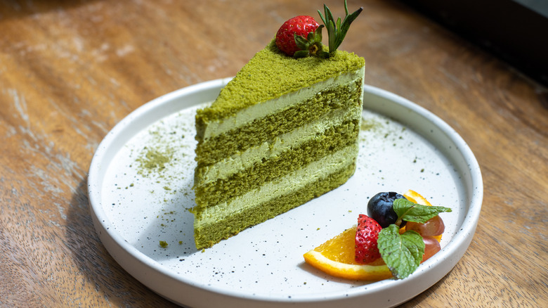 Slice of green matcha layer cake with a strawberry on top on a round white plate with a fruit garnish in front