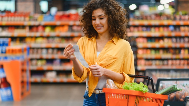 Person holding a shopping basket with groceries smiling while examining a long paper receipt in a retail store aisle