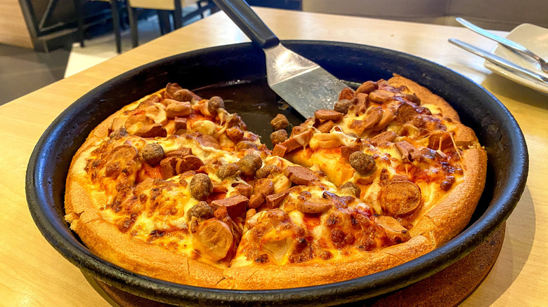 A PIzza Hut meat lovers in a pan on a table with a slice missing and serving spatula.