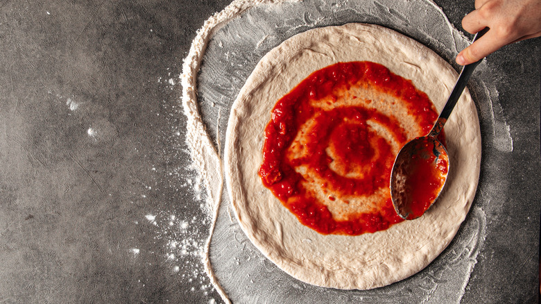 Top view of pizza sauce being spread on raw pizza dough with metal spoon