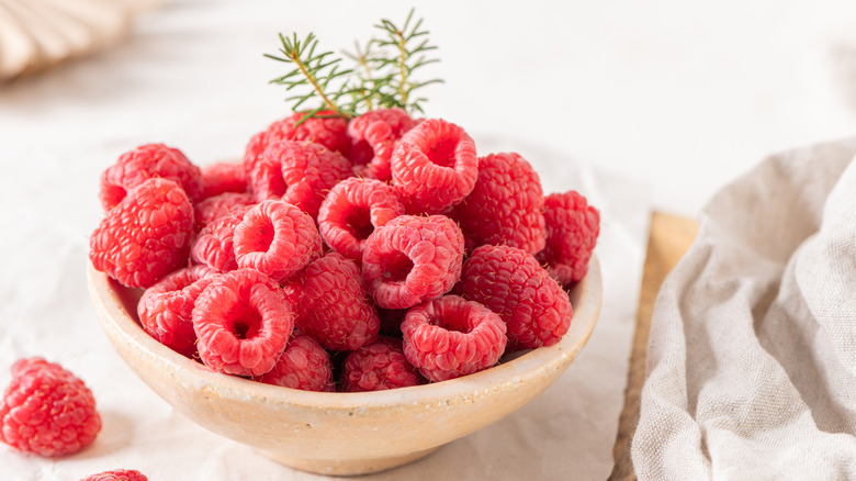 A rustic ceramic bowl filled with plump, fresh raspberries sits on a white table