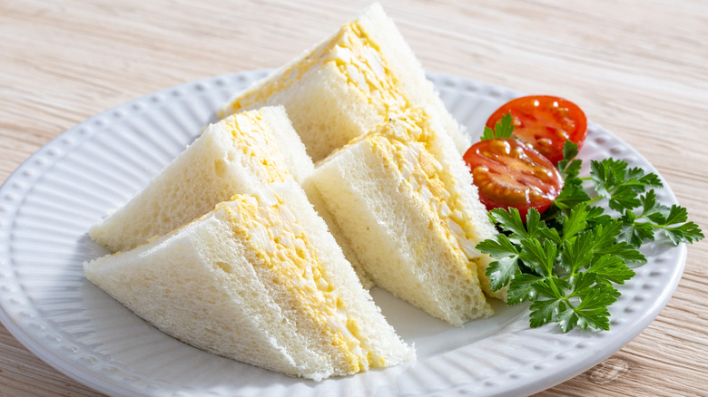 White plate with 4 egg salad sandwich triangles, tomato, and parsley.