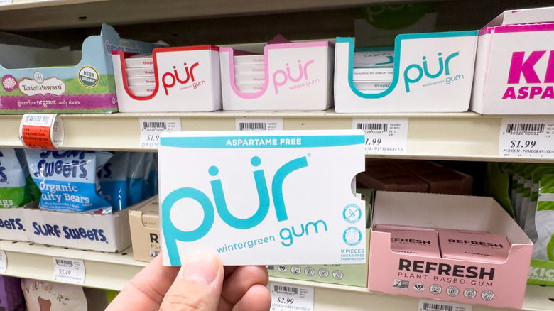 Pür gum in hand with packs of it behind on store shelves