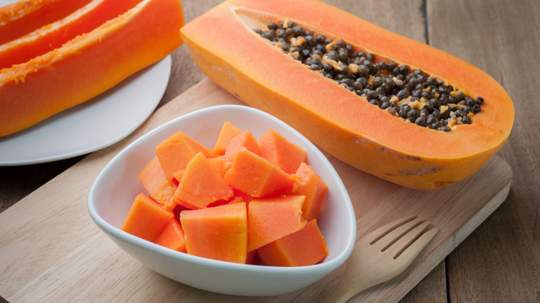 White bowl with chopped papaya next to half of a papaya with seeds inside -- sliced papaya on plate behind