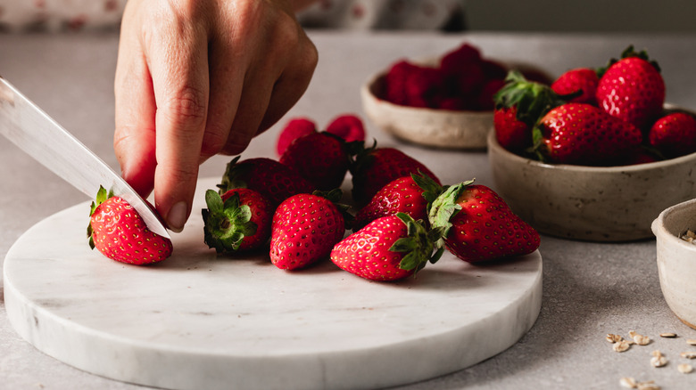 Person's hand holding strawberry as knife slices it in half on top of marble round with several strawberries to the right and in the background in another bowl