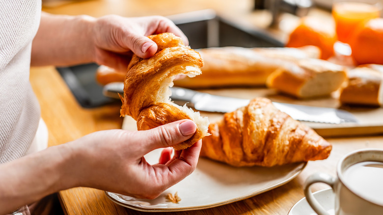 Hands pulling apart a croissant in a kitchen near the sink and a cup of coffee.