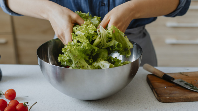 Two hands tossing lettuce leaves in a silver bowl in a kitchen