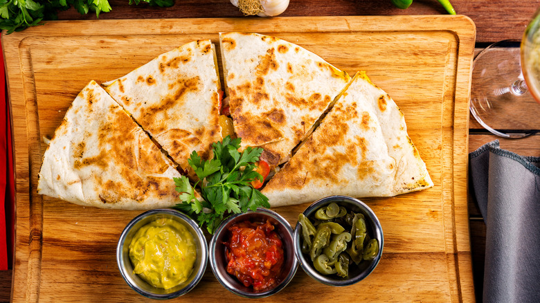 A cooked quesadilla on a wood cutting board with salsa, sauces, and peppers in ramekins.