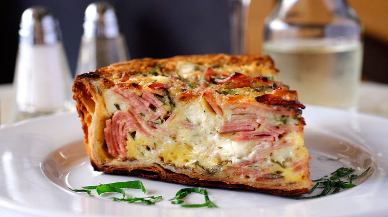Close up of a slice of ham and cheese quiche on a white plate, with salt and pepper shakers, and a water carafe blurred on a table in the background.