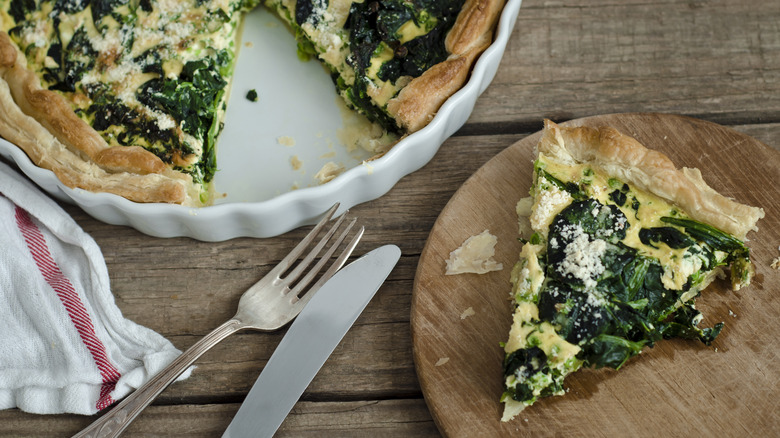 Aerial view of spinach quiche with a slice cut out and on a wooden plate nearby, along with utensils and a napkin.
