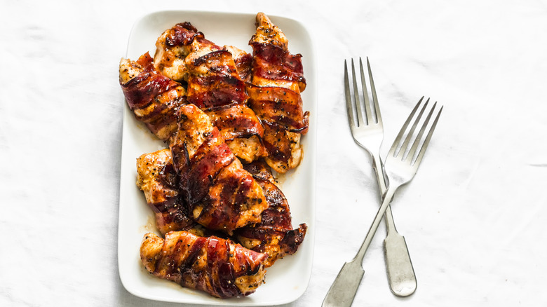 A top-down view of several bacon-wrapped chicken breasts on a white plate with two forks beside it.