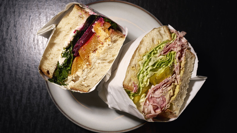 Halves of two wrapped gourmet sandwiches on a small white plate and dark table.