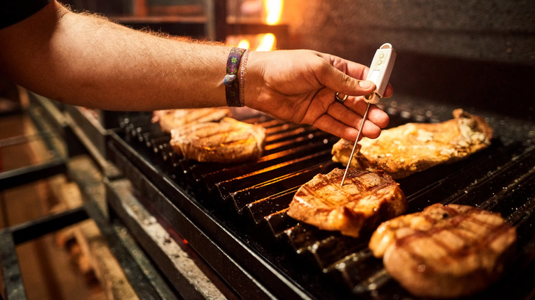 A hand sticking a meat thermometer into a pork steak as it cooks on a grill