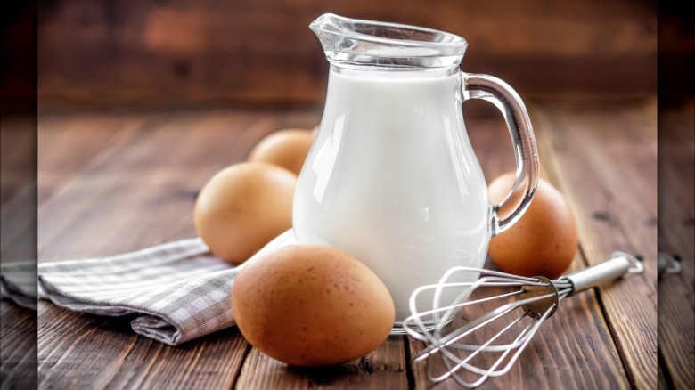 A pitcher of milk with several whole eggs around it with a whisk and farmhouse decor.