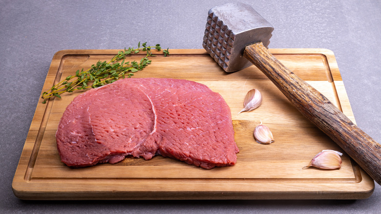 Piece of raw tenderized steak on cutting board next to a meat mallet