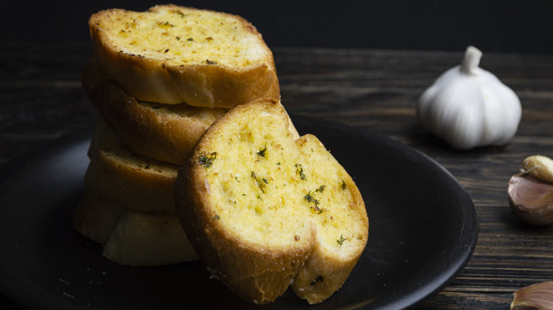 Slices of crispy garlic bread stacked on a black plate with a raw garlic bulb in the background.