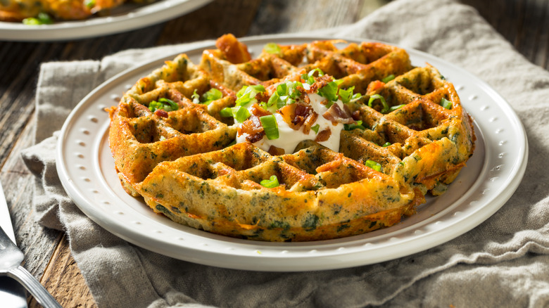 A savory egg waffle on a white plate with sour cream, green onions, and bacon on top