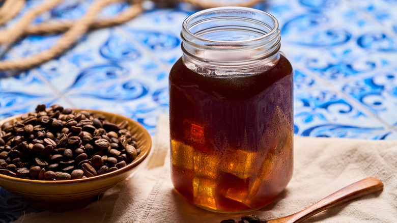 A mason jar of iced coffee rests on a tea towel, a bowl of coffee beans and a wooden spoon on either side.
