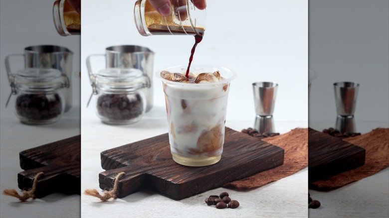 A hand pours a dark liquid sweetener from a measuring glass into a cup of a milky iced coffee on a wooden cutting board.