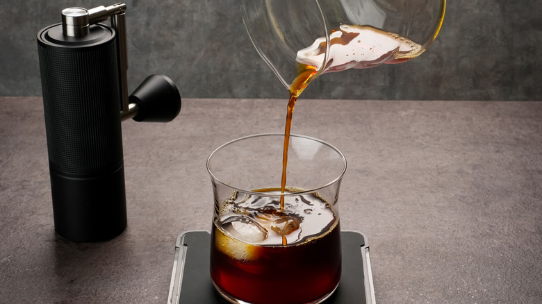 A jug of iced coffee is being poured into a glass. A black coffee grinder rests on the table in the background.