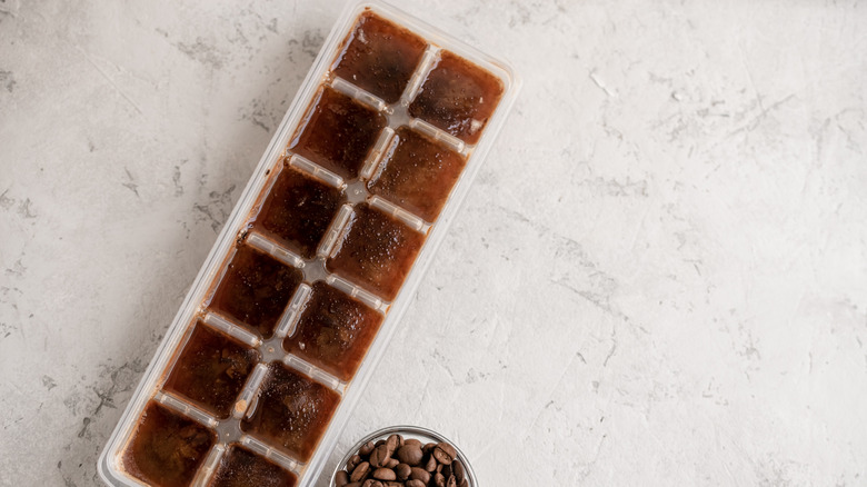 A transparent, plastic ice cube tray filled with frozen coffee cubes rests on a marbled table.