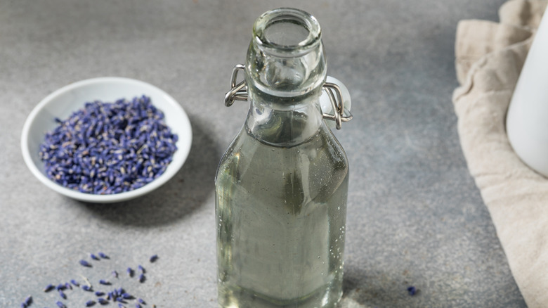 Homemade lavender simple syrup in a glass bottle on a table. Petals of lavender sit in a bowl to the left.
