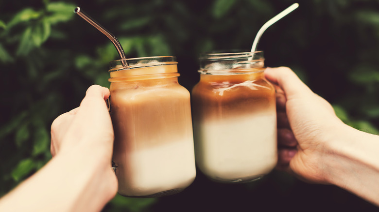 Two hands are clinking mason jars of iced coffee with milk, metal straws sticking out of both.