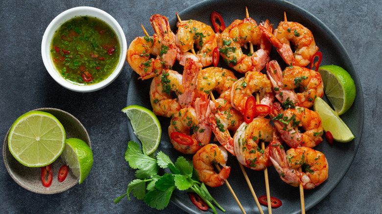 Four skewers of grilled shrimp on a black plate with lemon wedges, cilantro, and a small bowl of liquidy dip