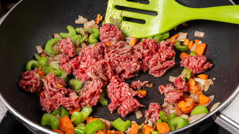 A skillet frying up ground beef and vegetables with a spatula nearby