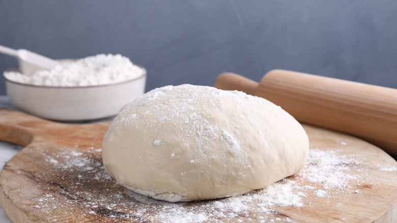 Ball of raw pizza dough on wooden board with flour and rolling pin in background.