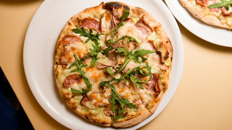 A personal pizza topped with ham and arugula on white plate