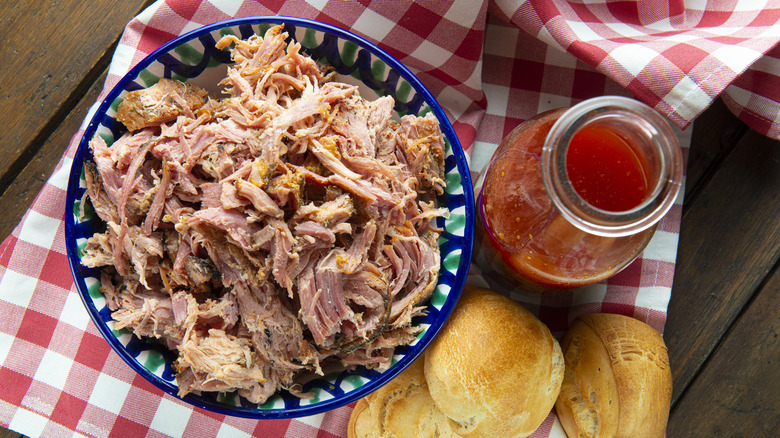 Pulled pork in bowl on wooden table alongside vinegar-based sauce and rolls.
