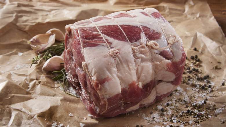 A trussed raw pork shoulder roast surrounded by herbs and spices sitting on butcher paper